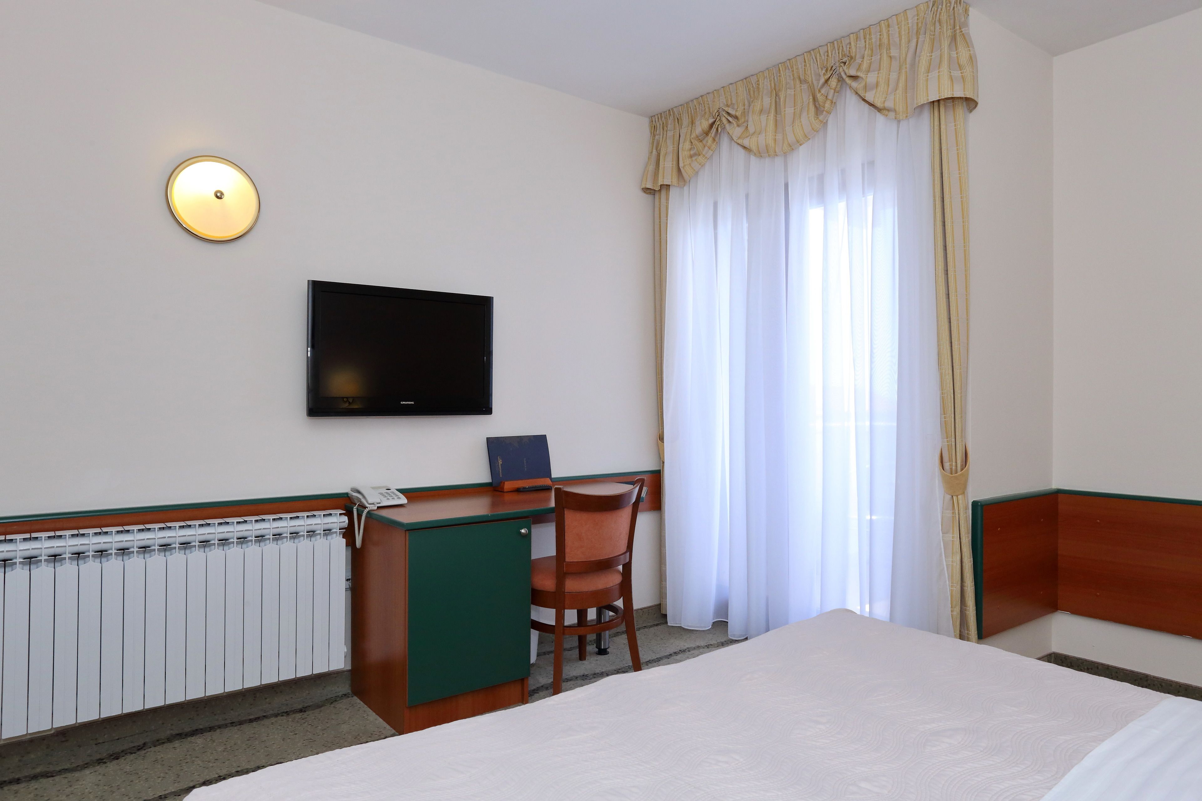 Una camera d'albergo con un letto, una scrivania con una sedia, una televisione a parete e una finestra con tende.