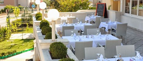 Un área de comedor al aire libre con mesas dispuestas para dos personas, rodeada de vegetación y vistas a la calle.