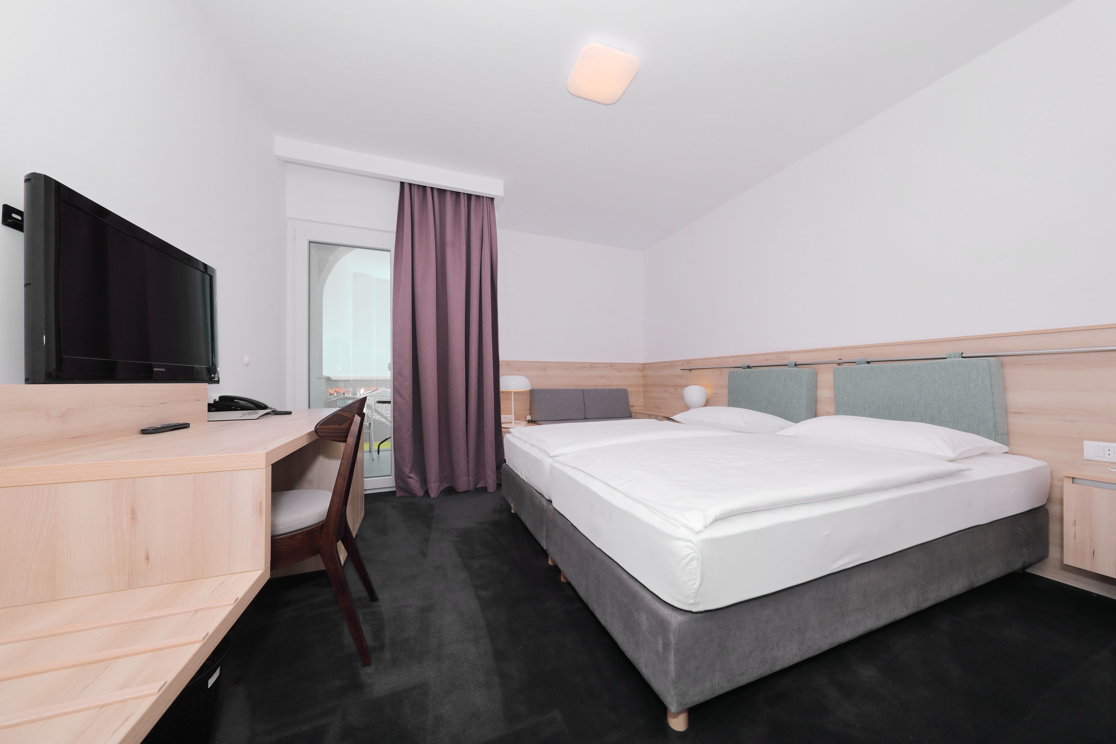 A hotel room with a bed, a desk, a chair, a TV, and a window with curtains.