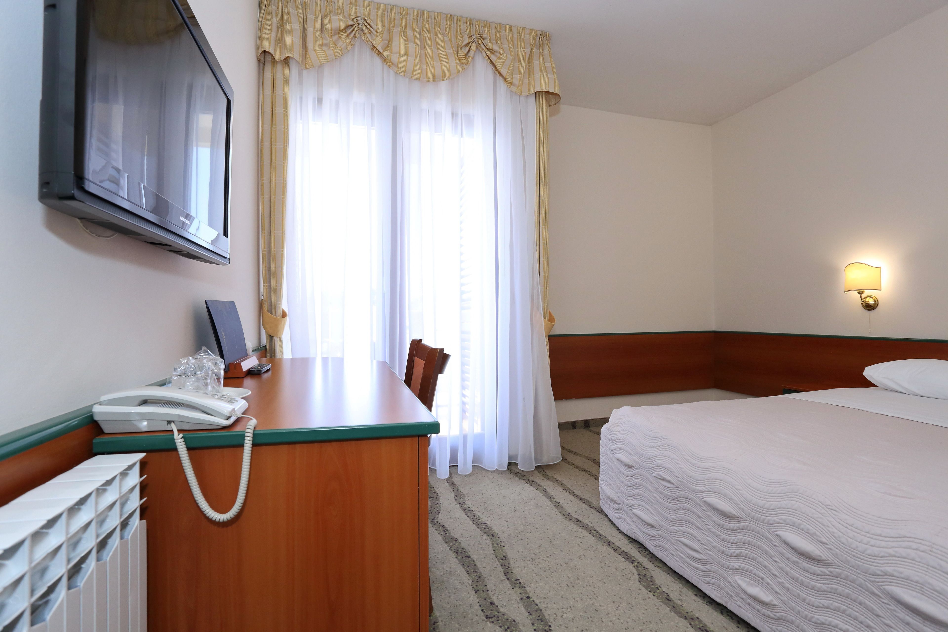 A hotel room with a bed, a desk with a telephone, a television mounted on the wall, and a window with curtains.