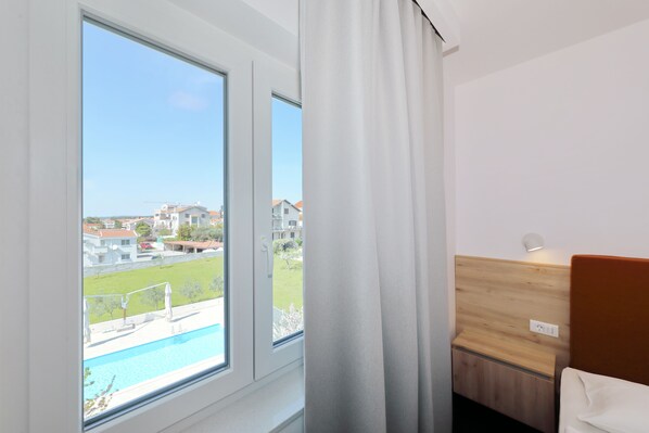 Une chambre d’hôtel dotée d’une grande fenêtre, d’une tête de lit en bois et offrant une vue sur une piscine et les bâtiments environnants.