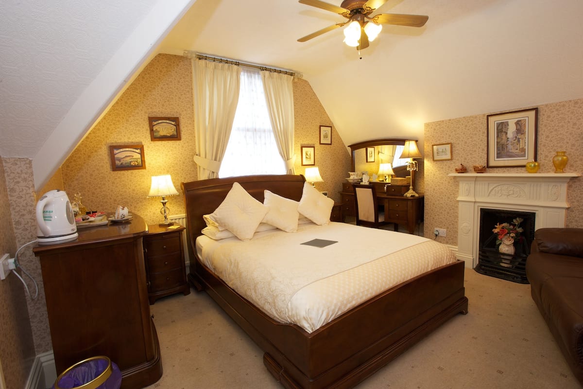 A bedroom with a large bed, a ceiling fan, a fireplace, and a nightstand with a lamp.
