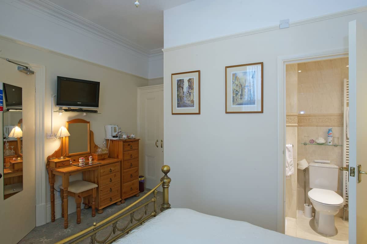 A hotel room with a wooden dresser, a television mounted above it, a bed with a white headboard, and a bathroom with a toilet and shower.