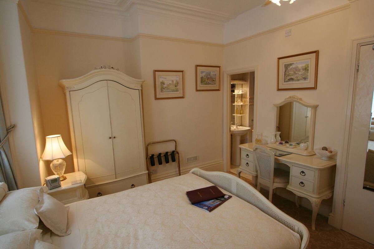 A hotel room with a large bed, a wardrobe, a vanity with a mirror, and a bedside table with a lamp.