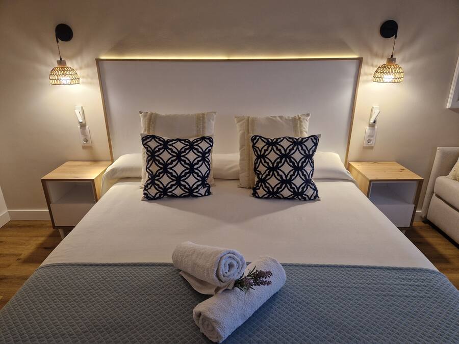 Una cama bien hecha con dos almohadones estampados, dos mesitas de noche y dos lámparas de pared.