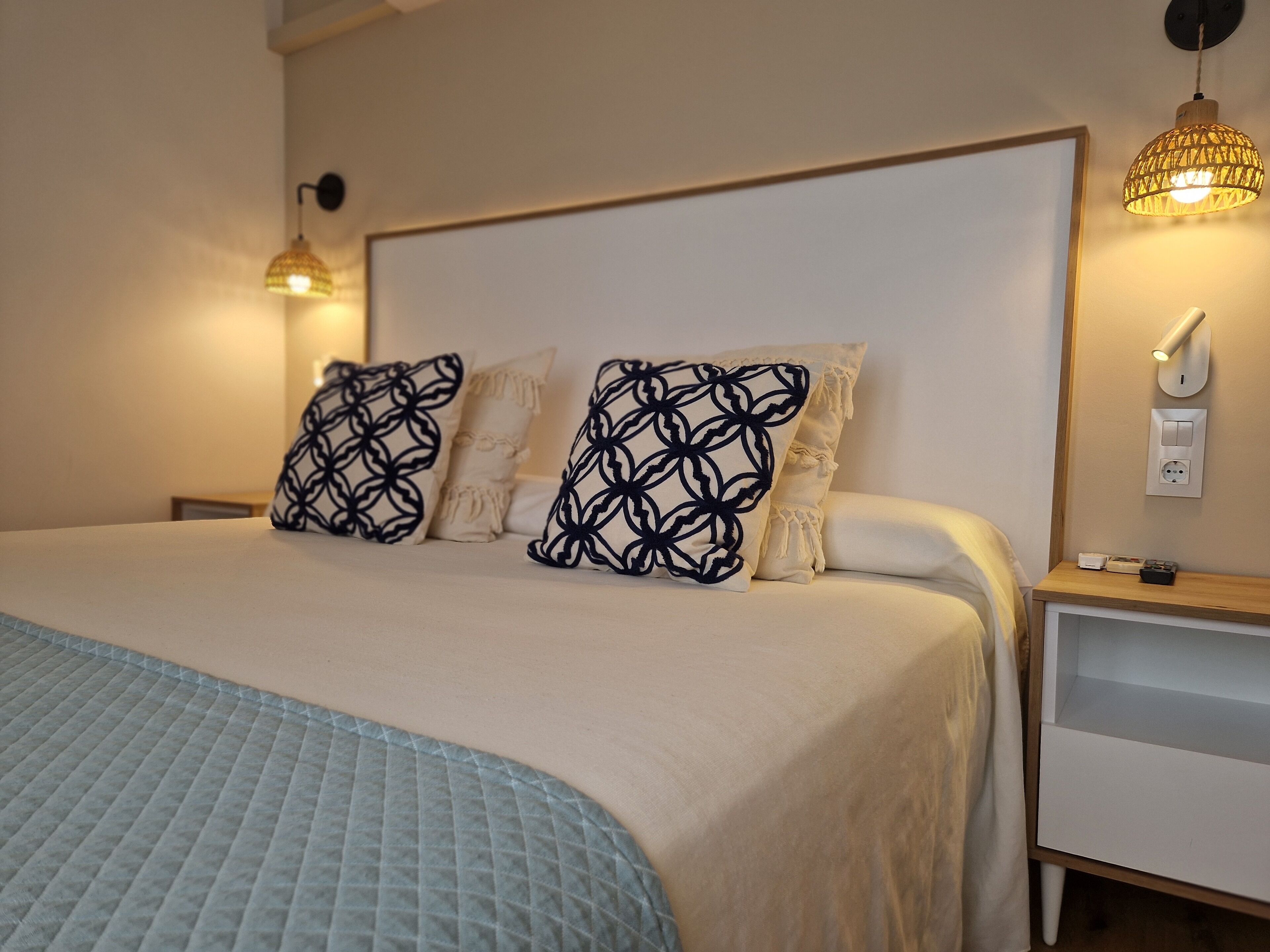 Ein ordentlich bezogenes Bett mit gemusterten Kissen, ein Nachttisch mit Lampe und eine Steckdose an der Wand.