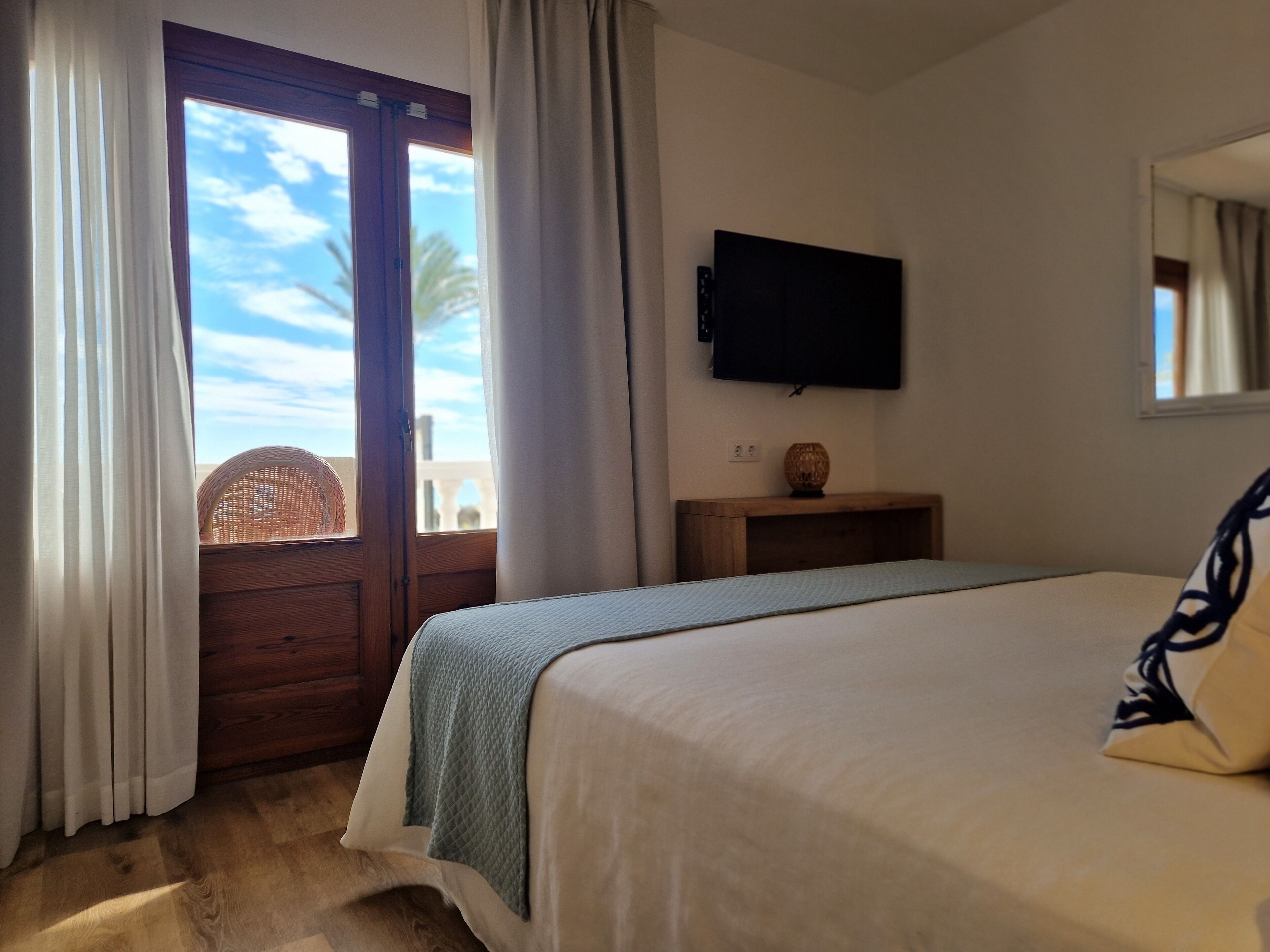A bedroom with a bed, a television mounted on the wall, a wooden nightstand, and a view of palm trees through a window.