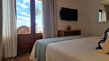 A bedroom with a bed, a television mounted on the wall, a wooden nightstand, and a view of palm trees through a window.