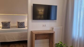 Una sala moderna con un banco de madera, un televisor de pantalla plana montado en la pared, una planta en maceta sobre una mesa de centro y una cama con almohadas estampadas.