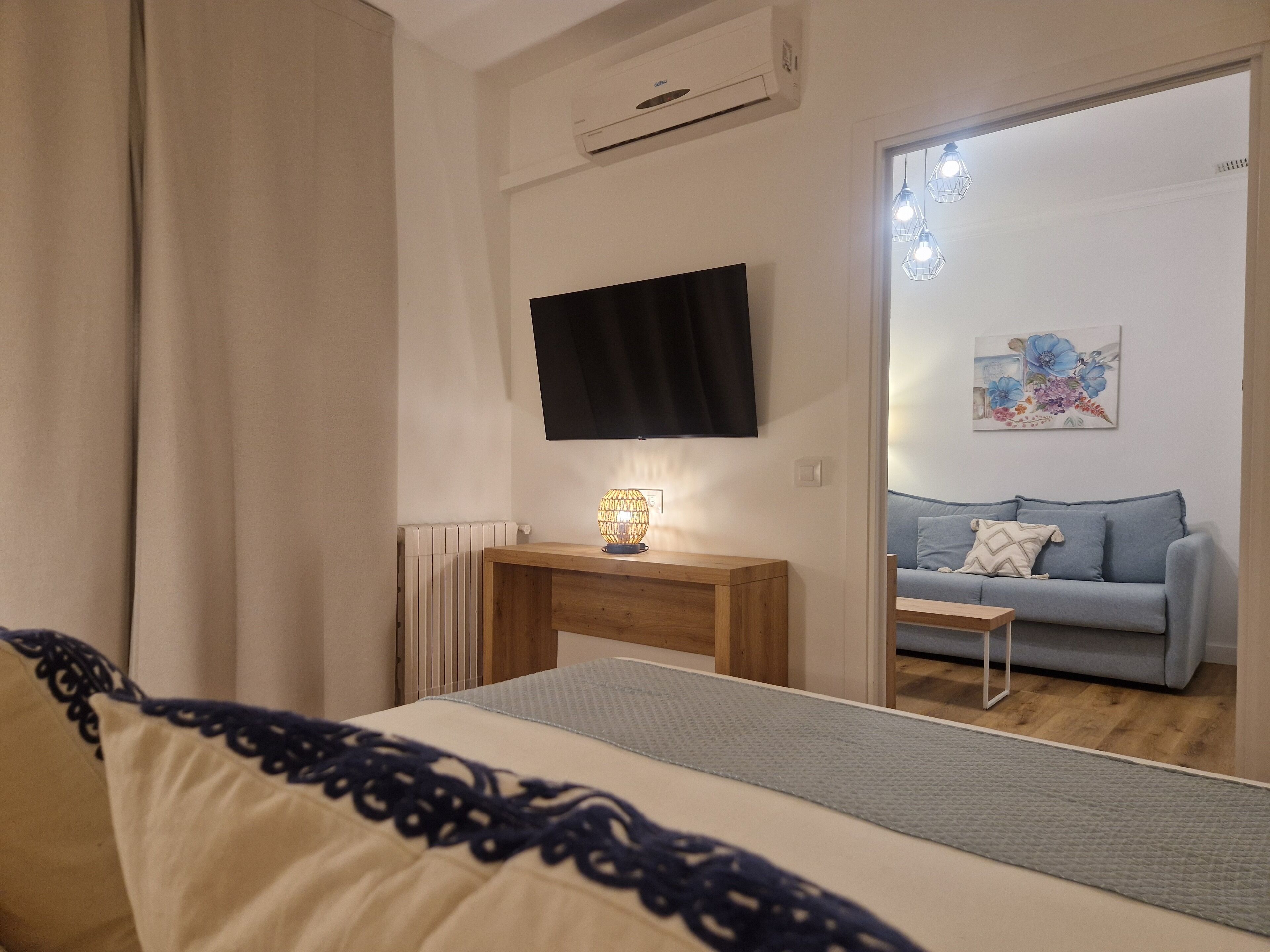 A bedroom with a bed, a bedside table with a lamp, a TV mounted on the wall, a radiator, and a view into another room with a sofa and a painting.