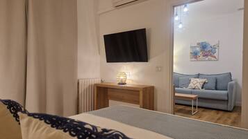 A bedroom with a bed, a bedside table with a lamp, a TV mounted on the wall, a radiator, and a view into another room with a sofa and a painting.