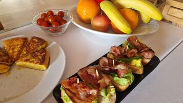 Una mesa de desayuno con tostadas, tocino, lechuga y tomate en un plato, y un bol de tomates cherry y una fuente de fruta en otro.