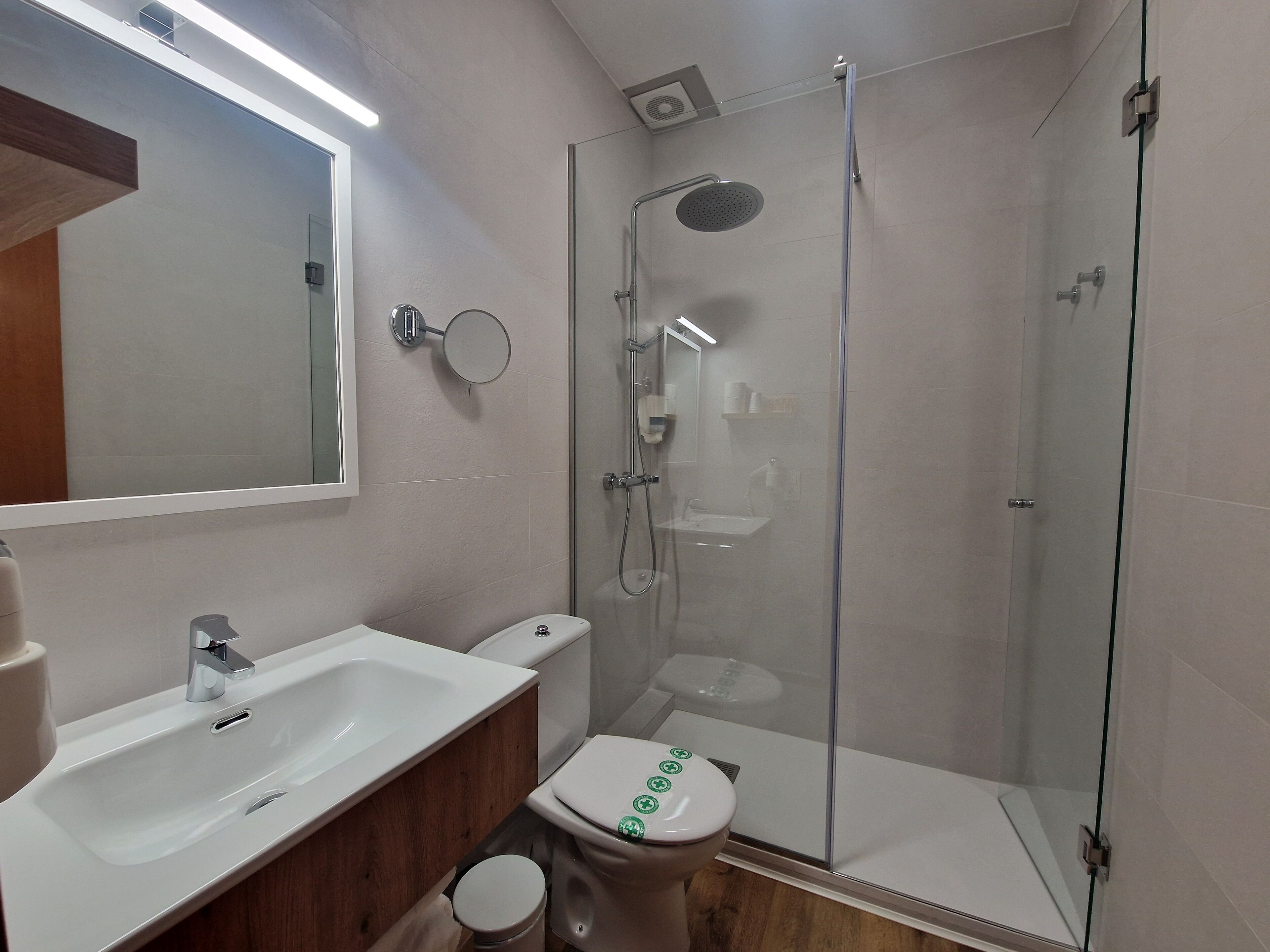 A modern bathroom with a glass-enclosed shower, a white sink, and a toilet with green symbols.