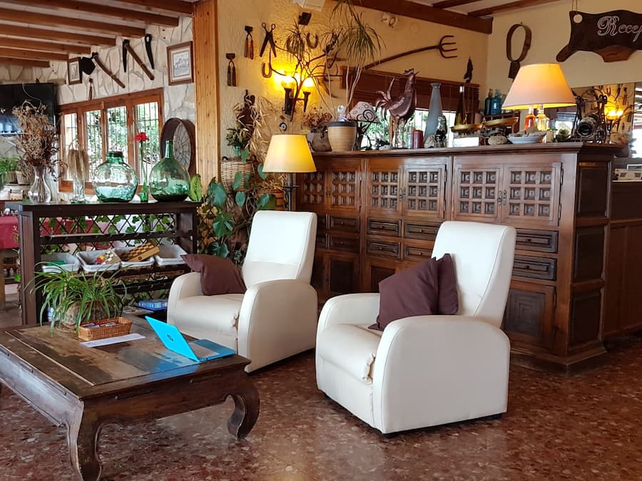 Un vestíbulo acogedor con una mesa de recepción, dos sillones blancos de cuero, una mesa de centro de madera con una computadora portátil y una pared decorativa con utensilios de cocina.
