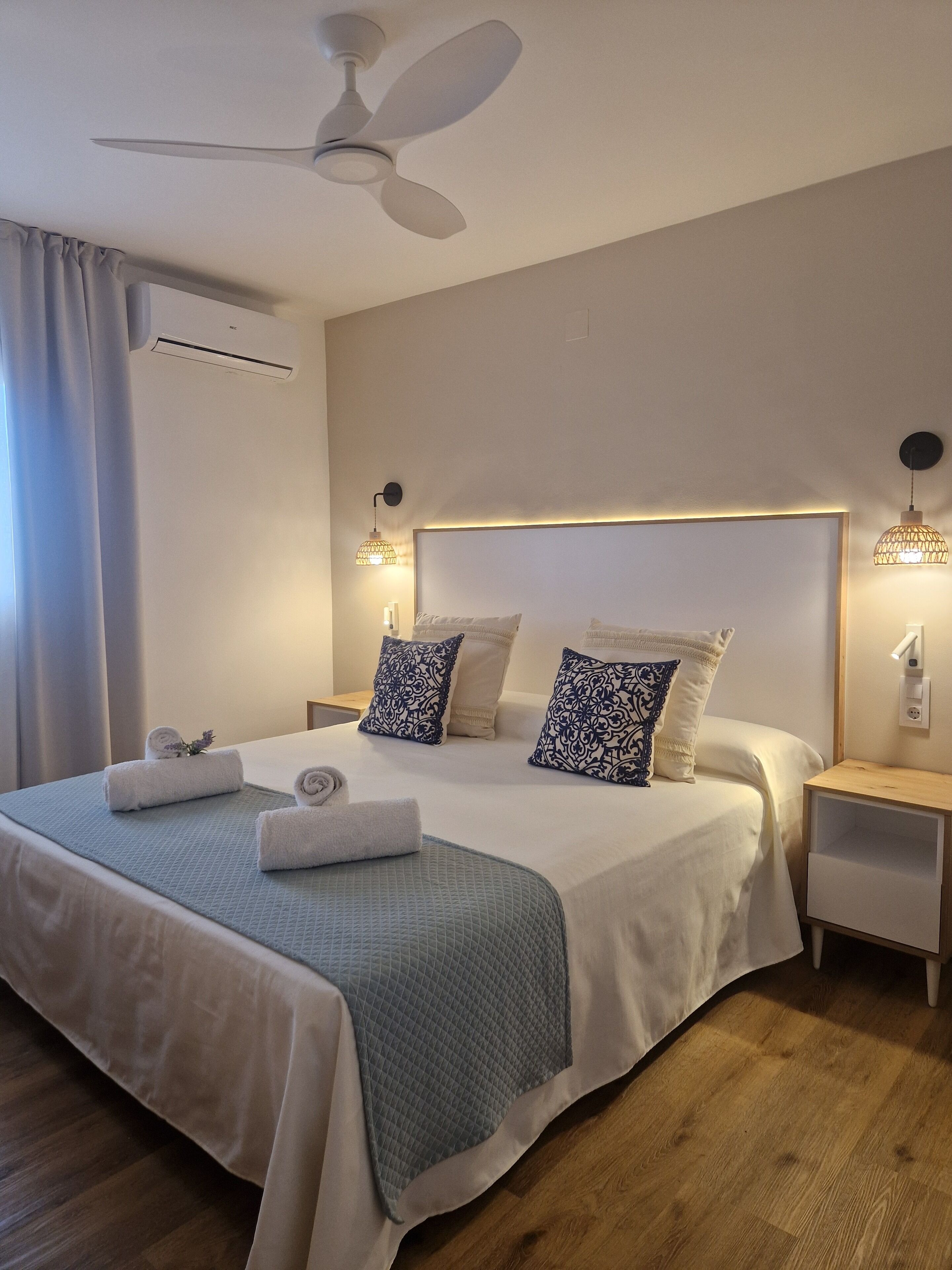 Ein ordentlich bezogenes Bett mit einer gesteppten Tagesdecke, zwei gemusterten Kissen und aufgerollten Handtüchern. An der Decke hängt ein Ventilator, an den Nachttischen stehen Lampen und an der Wand ist eine Klimaanlage angebracht.