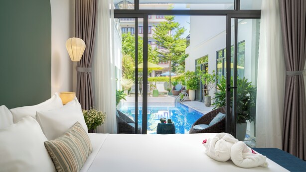 A hotel room with a bed, a view of a swimming pool, and a balcony with outdoor furniture. - Volar Hoi An (Da Nang)