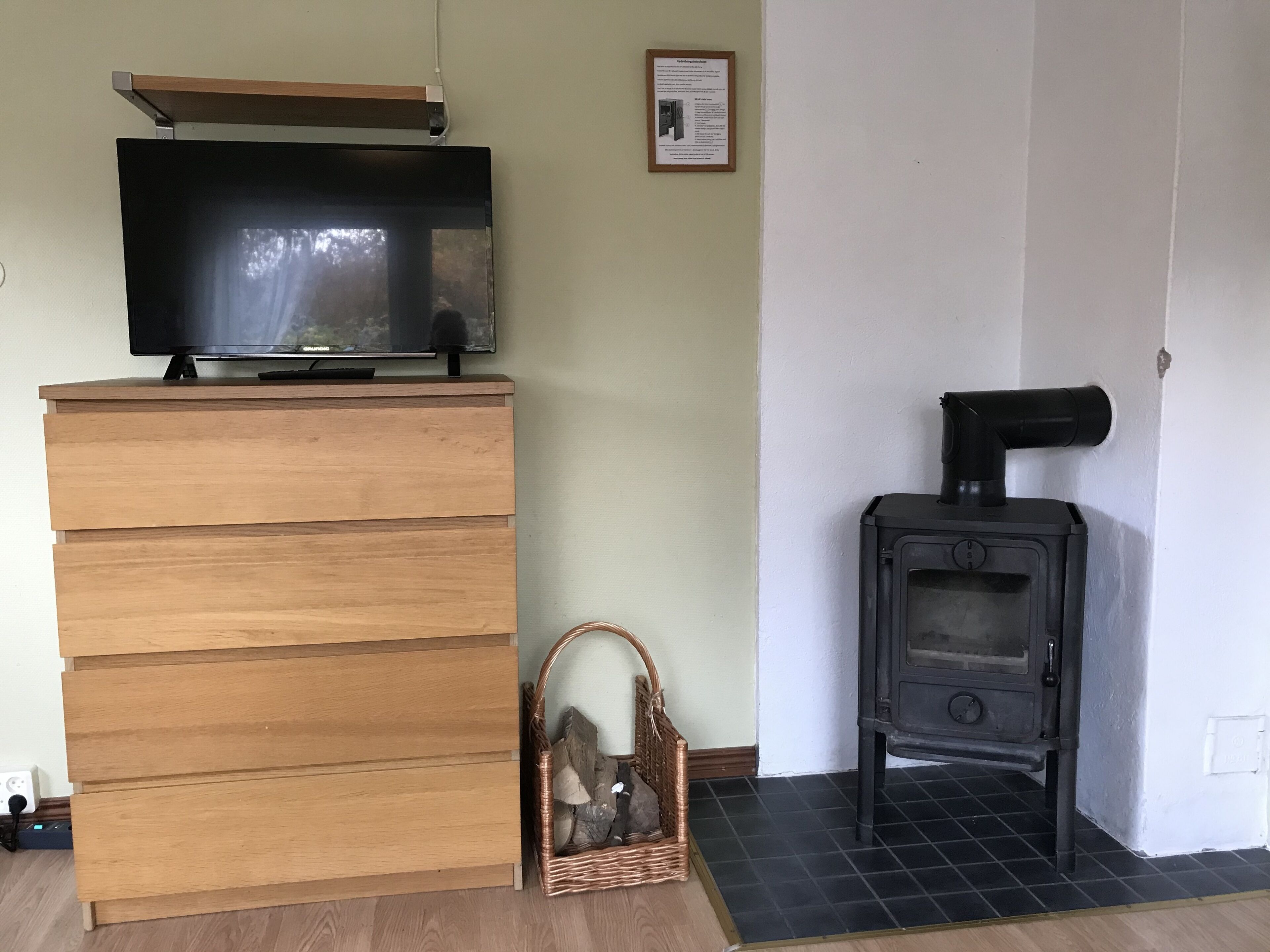 A room with a wooden dresser, a television on top, a wicker basket with firewood, and a black wood-burning stove.