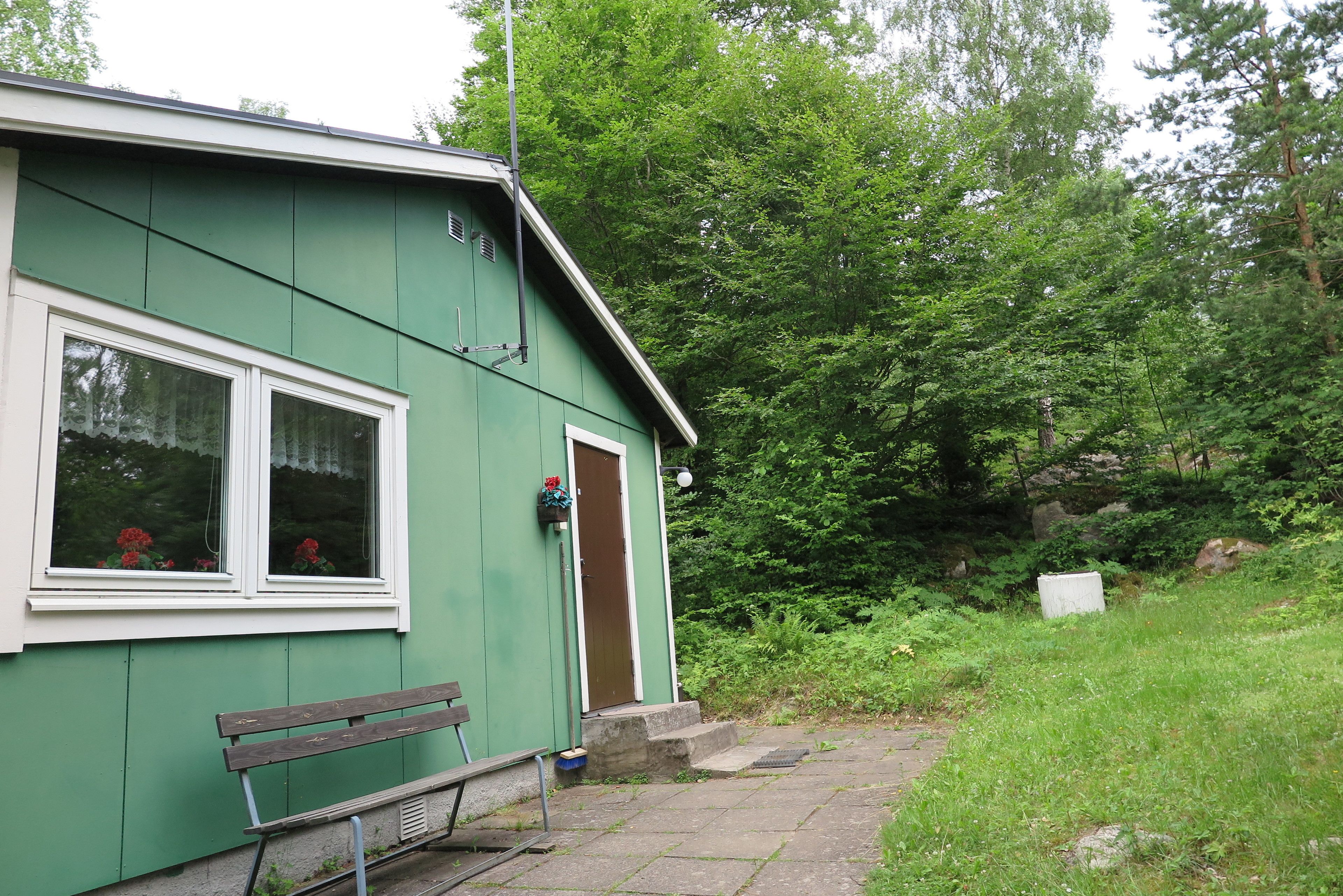 A green building with a bench outside, a door, and a window with red flowers.