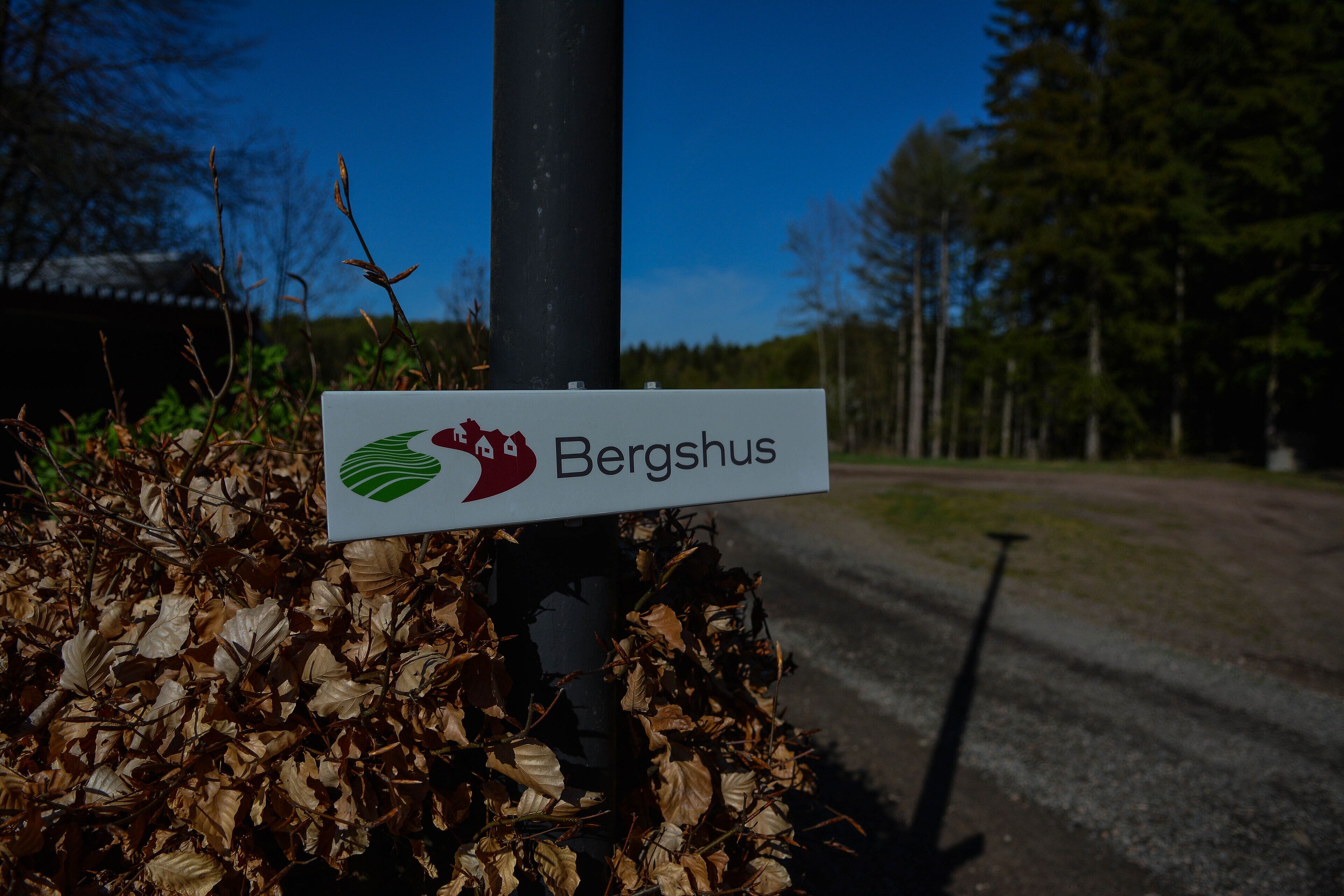 Ein Schild mit dem Namen "Bergshus" und einem Logo, an einem Pfahl angebracht, umgeben von trockenen Blättern.