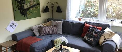 Uma sala de estar com um sofá cinza escuro, uma pequena mesa de madeira com um vaso de flores, uma parede verde com uma moldura e uma janela com vista para árvores.