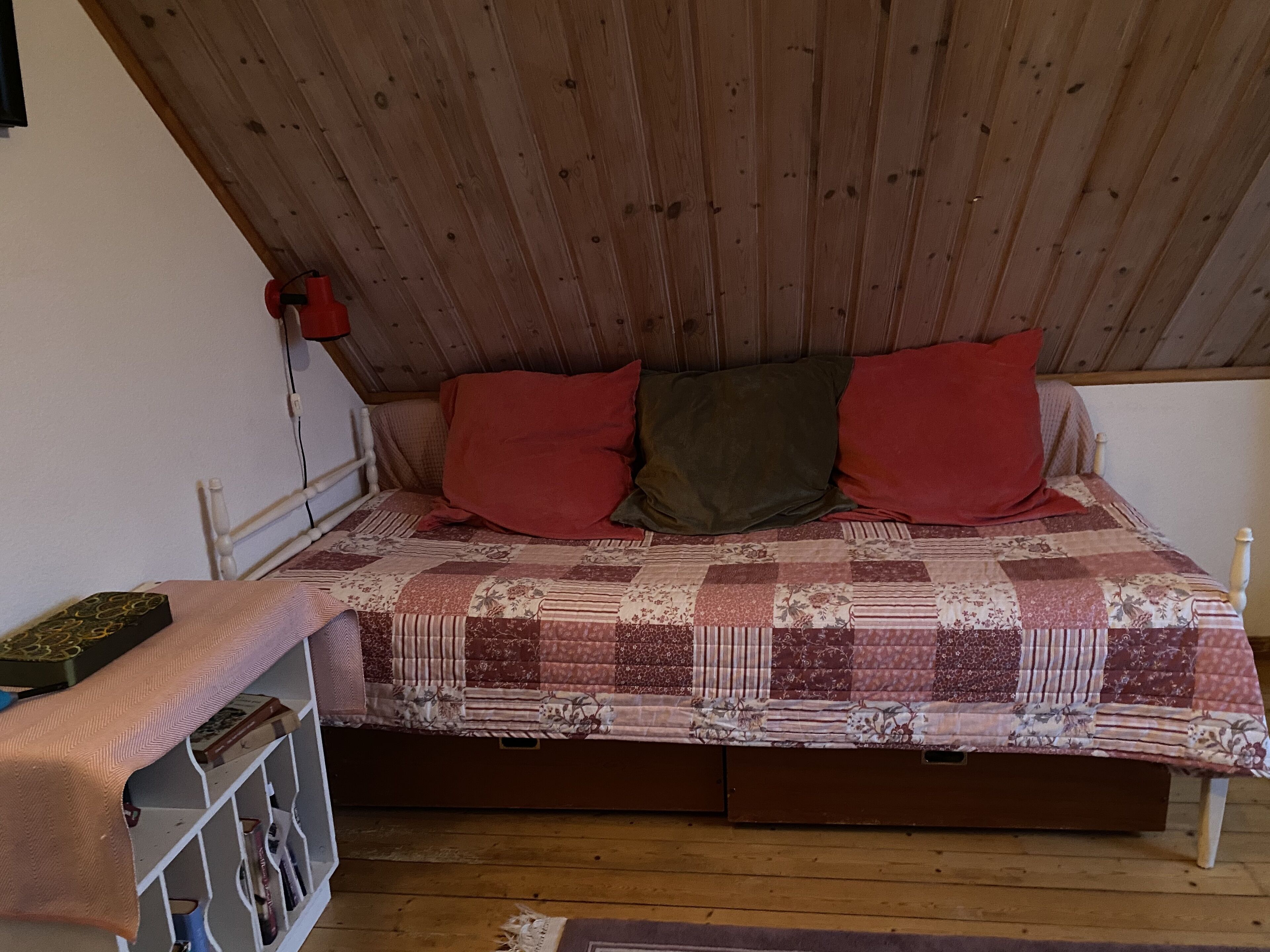 Habitación individual con techo de madera, colcha acolchada y mesita de noche con libros y una lámpara.
