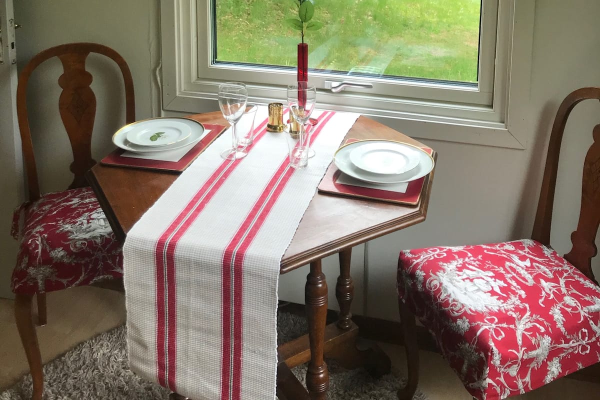 A dining area with a table set for two, a window with a view of greenery, and a red and white patterned curtain.
