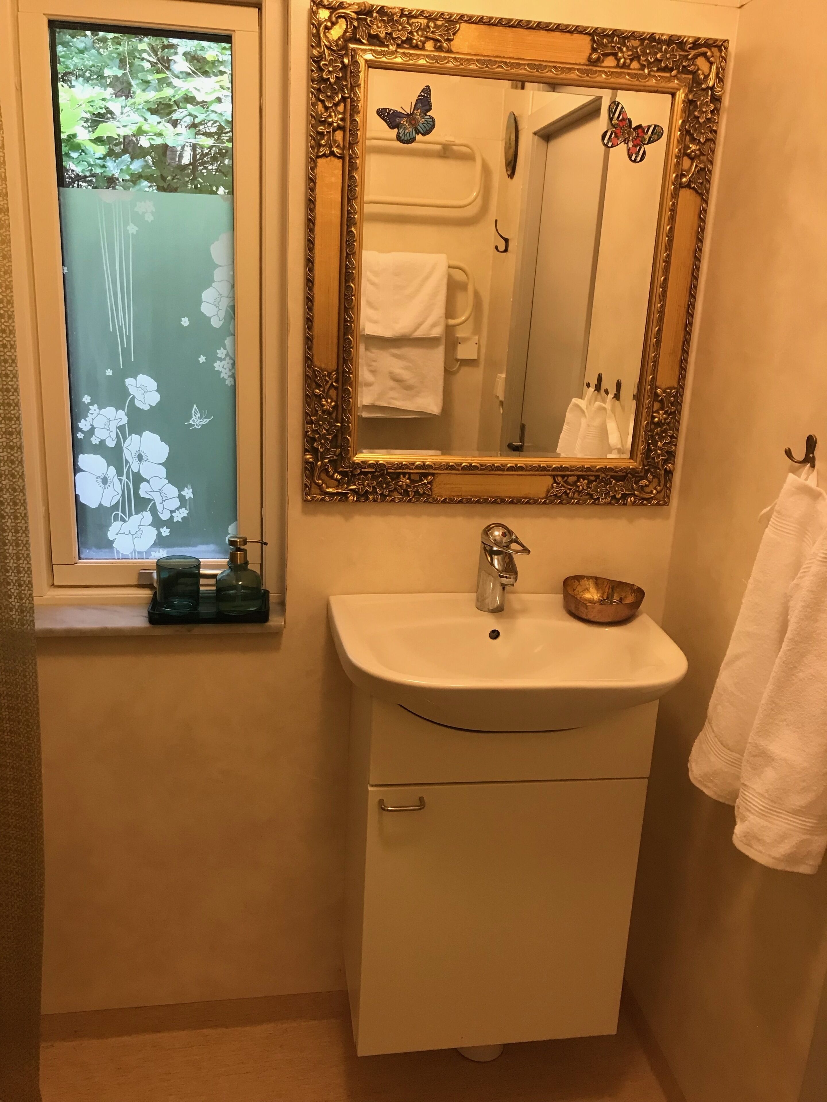 Un bany amb un lavabo blanc, un mirall amb marc daurat i una finestra amb un disseny floral.