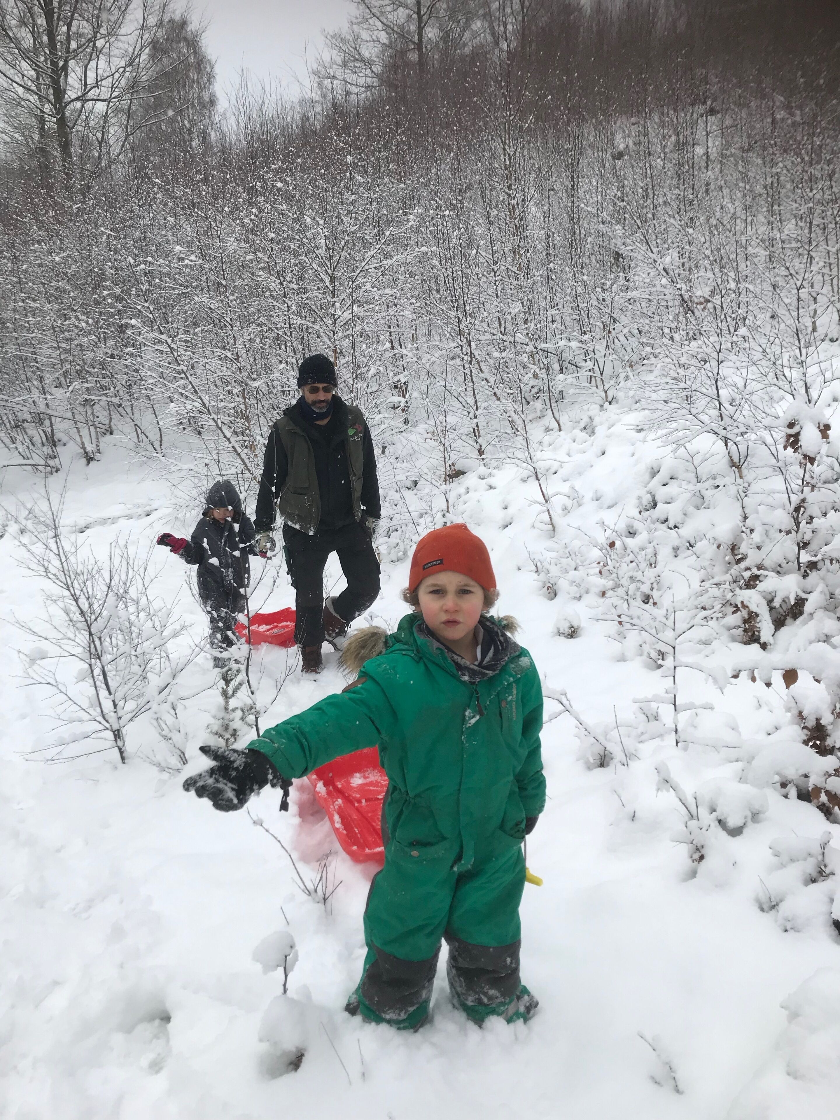 A snowy outdoor scene with a person in green winter clothing and a child in a red hat.