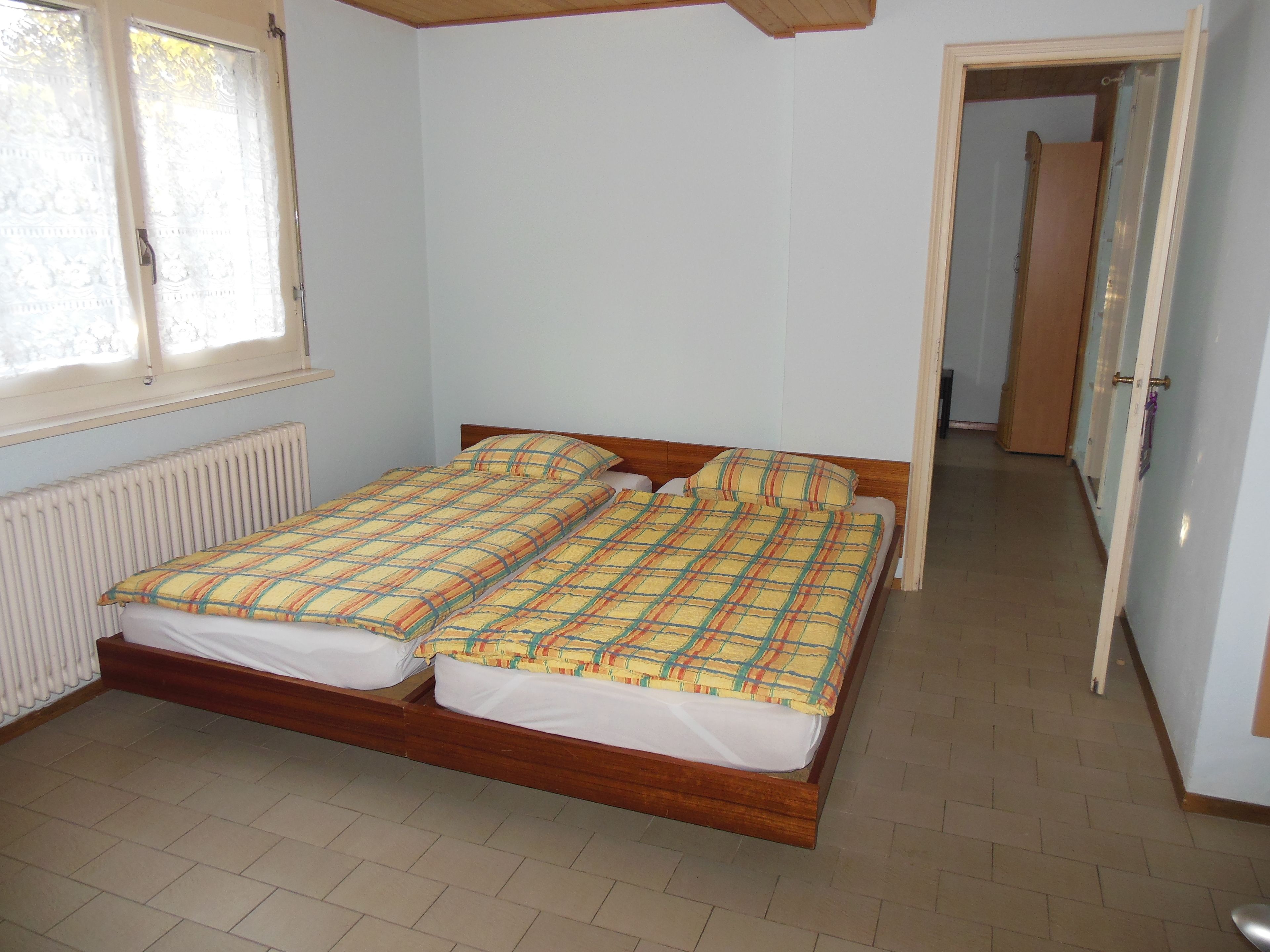 Una habitación con una cama de madera, paredes blancas, un radiador y suelo de baldosas.