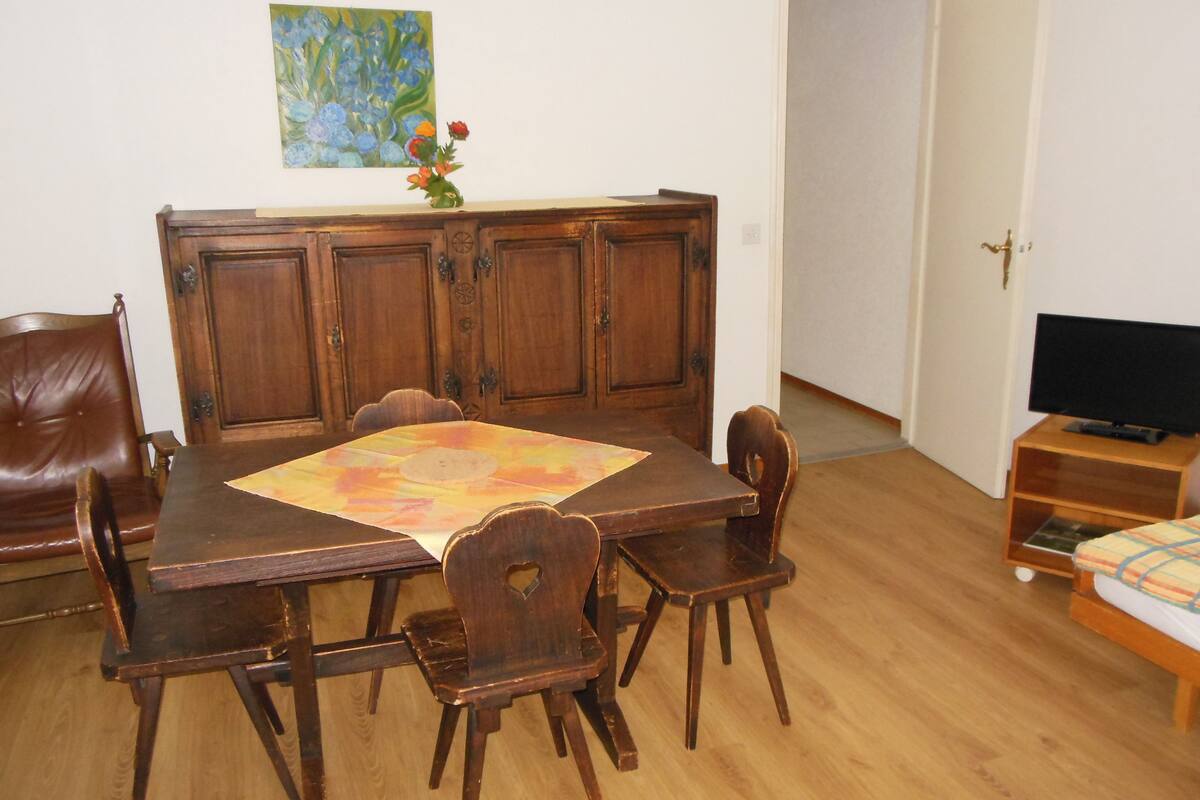 Un coin repas avec une table en bois, des chaises et un buffet. Il y a un tableau au mur et un téléviseur posé sur un meuble.