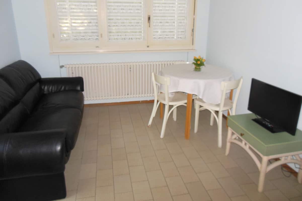 Une pièce avec un canapé en cuir noir, une petite table ronde recouverte d’une nappe blanche, une table verte et un téléviseur noir posé sur un meuble.