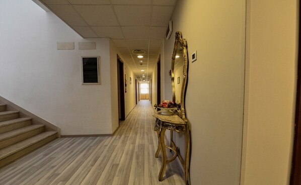 Un couloir d’hôtel avec un escalier à gauche, un miroir à droite et une porte au fond.