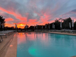 Une piscine entourée d’arbres, avec des chaises longues et des parasols, au moment du coucher du soleil.
