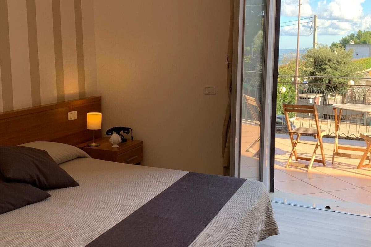 A hotel room with a bed, bedside table with a lamp, and a view of the outside balcony.
