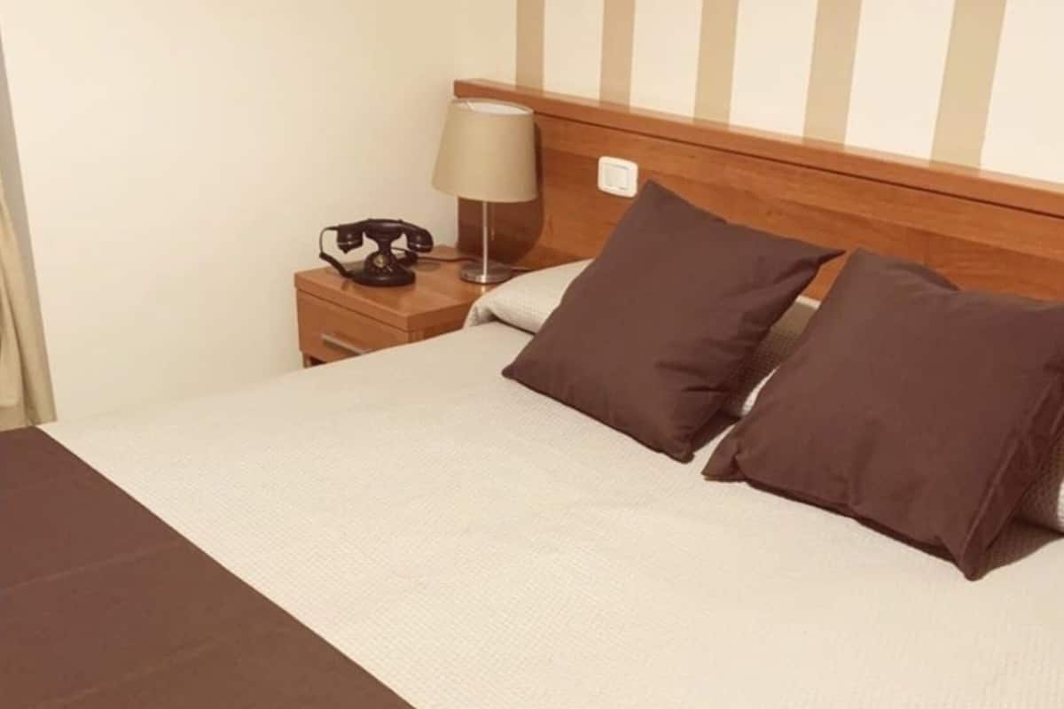 A neatly made bed with a brown bedspread and two pillows, a bedside lamp, and a telephone on a wooden nightstand.