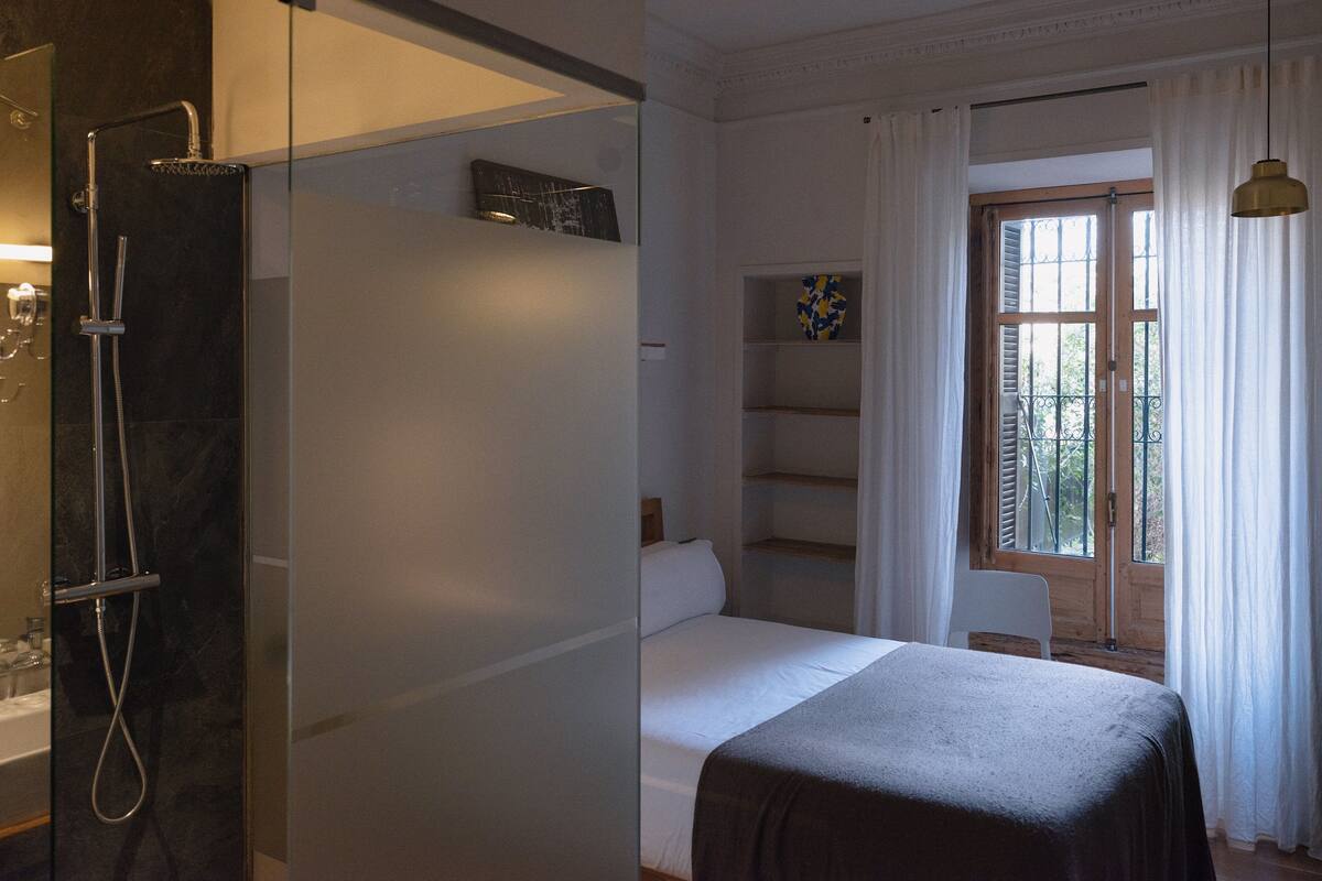 A hotel room with a shower, a bed, a window with curtains, and a shelf with decorative items.