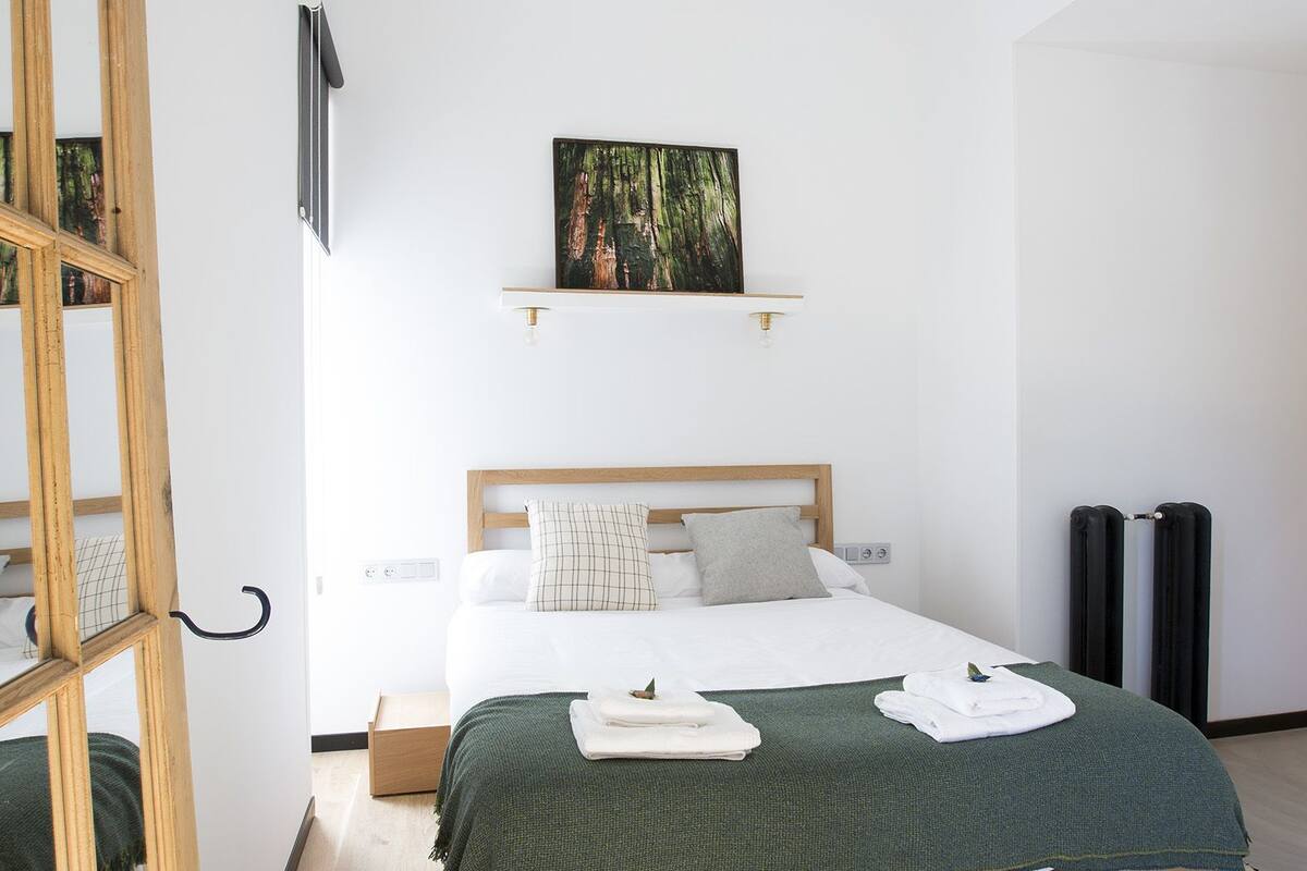 A modern bedroom with a wooden bed, a green throw blanket, a hanging gold pendant light, and a painting on the wall.