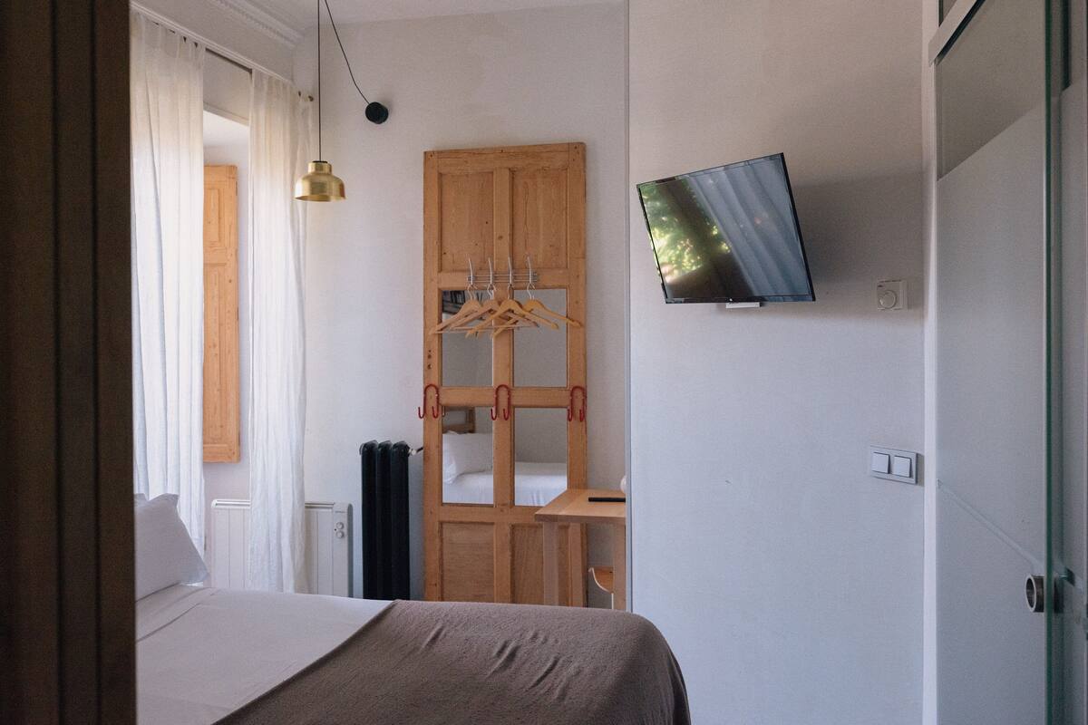 A bedroom with a bed, a wooden wardrobe, a television mounted on the wall, and a hanging light fixture.