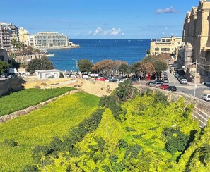 Un paisaje urbano costero con vista al mar, una calle con coches aparcados y edificios al fondo.