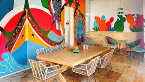 Un comedor con una mesa de madera, sillas de mimbre y un mural colorido en la pared.