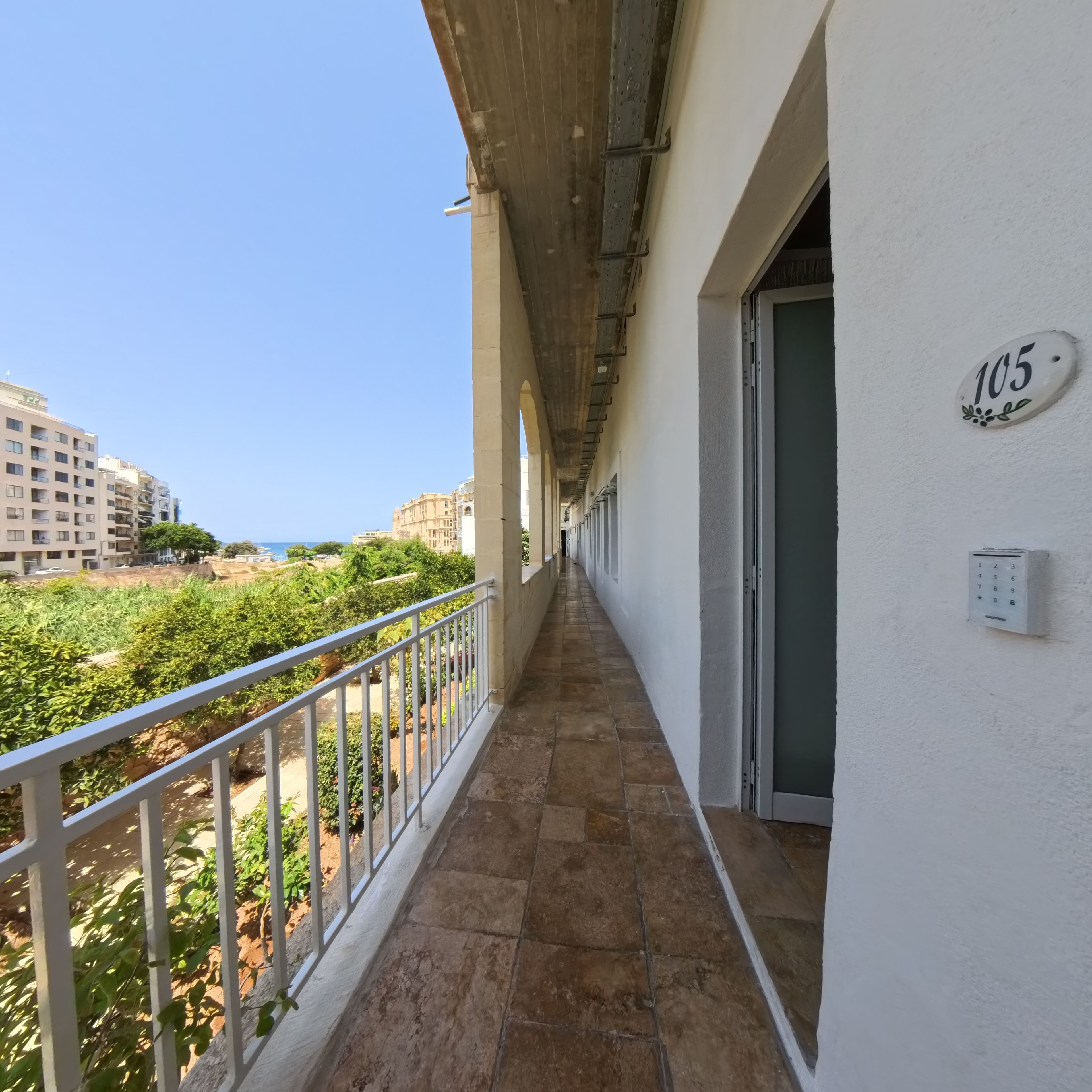 A corridor with a balcony, a door marked with the number 105, and a view of buildings and greenery.