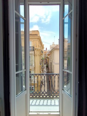 Blick von einem Balkon mit schmiedeeisernem Geländer auf eine schmale Straße gesäumt von Gebäuden mit klassischer Architektur und einem wolkenlosen Himmel.