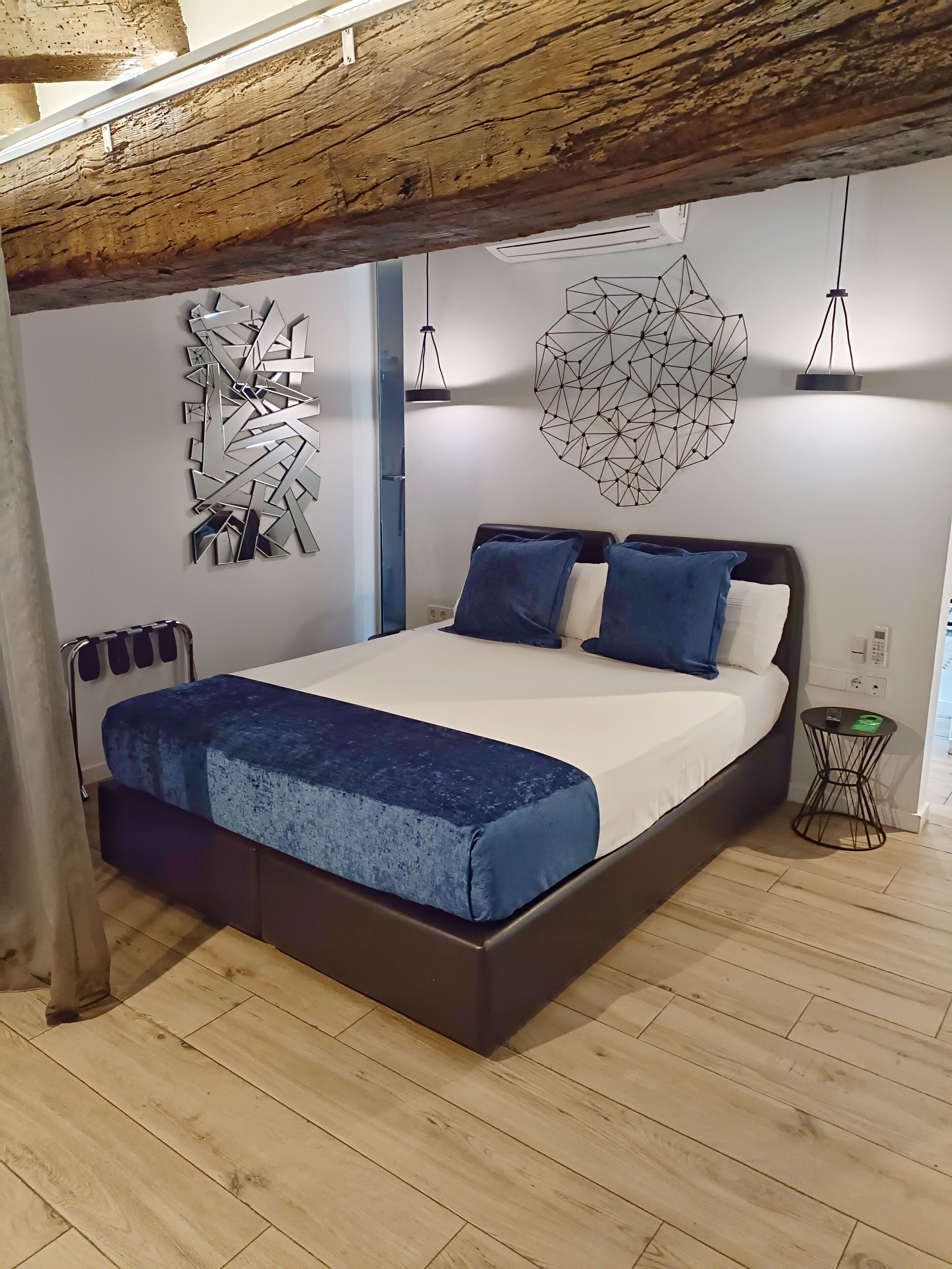 A modern bedroom with a wooden ceiling, a bed with a blue bedspread, two blue pillows, and geometric wall art.