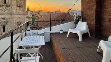 Terraza en la azotea con mobiliario de plástico blanco, un muelle de madera y vistas a una torre histórica y al paisaje urbano al atardecer.