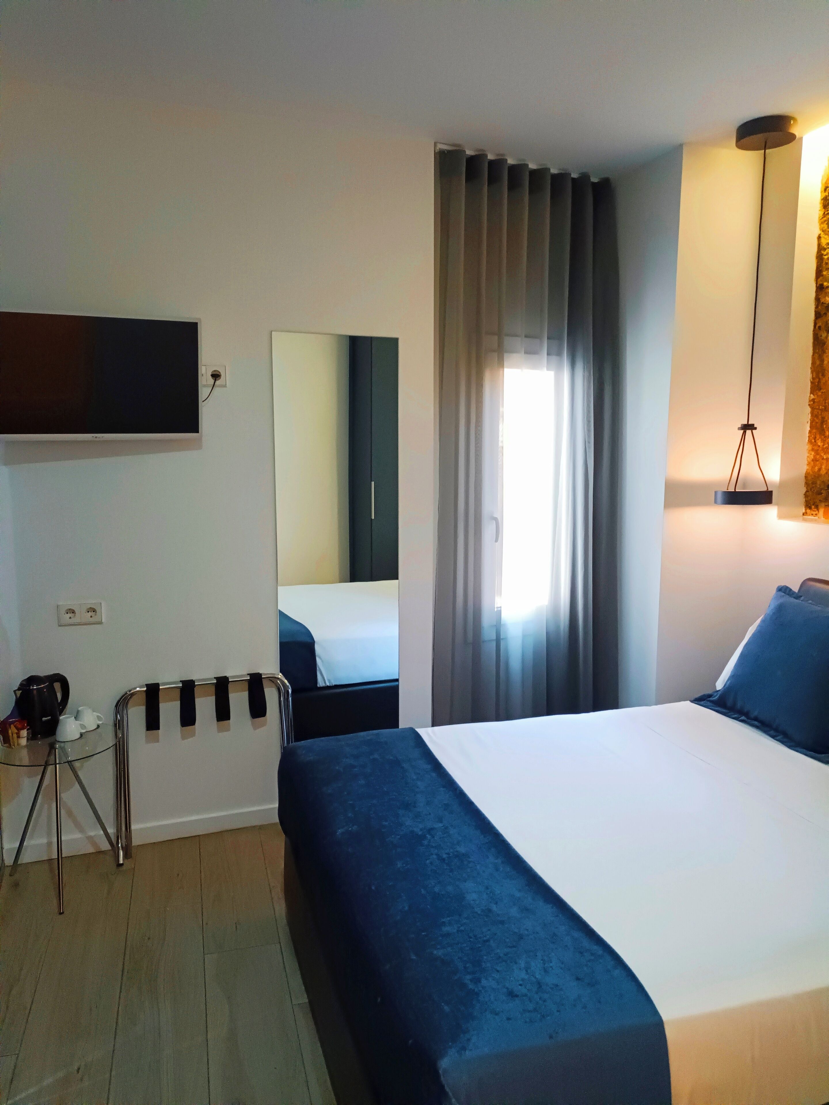A hotel room with a large bed, a TV mounted on the wall, a small table with a kettle and cups, and a mirror reflecting the room.