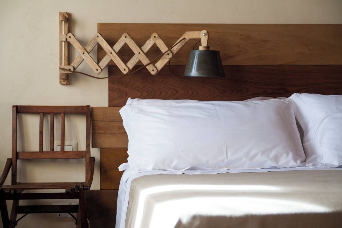 Una cama con sábanas blancas, cabecera de madera y una lámpara fijada a la pared.
