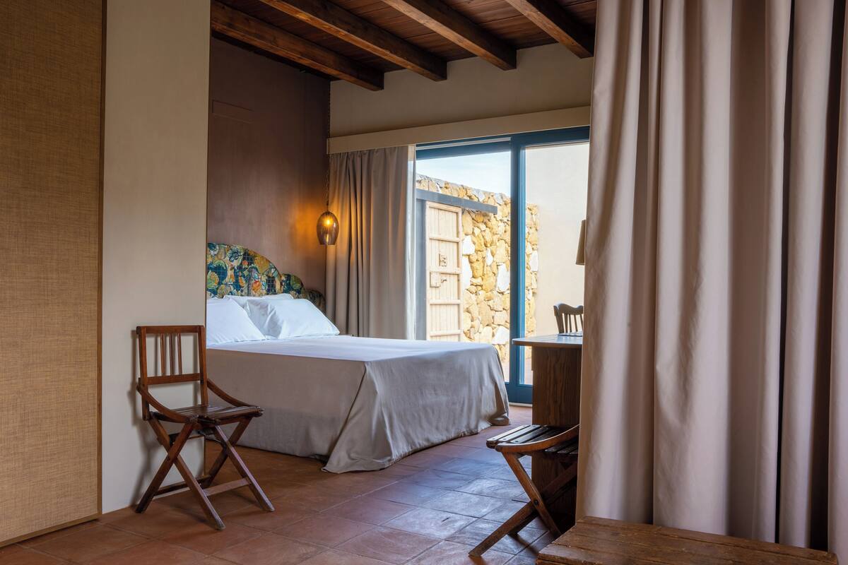 Una habitación con una cama, dos sillas de madera, una mesa, un muro de piedra y una ventana con cortinas.
