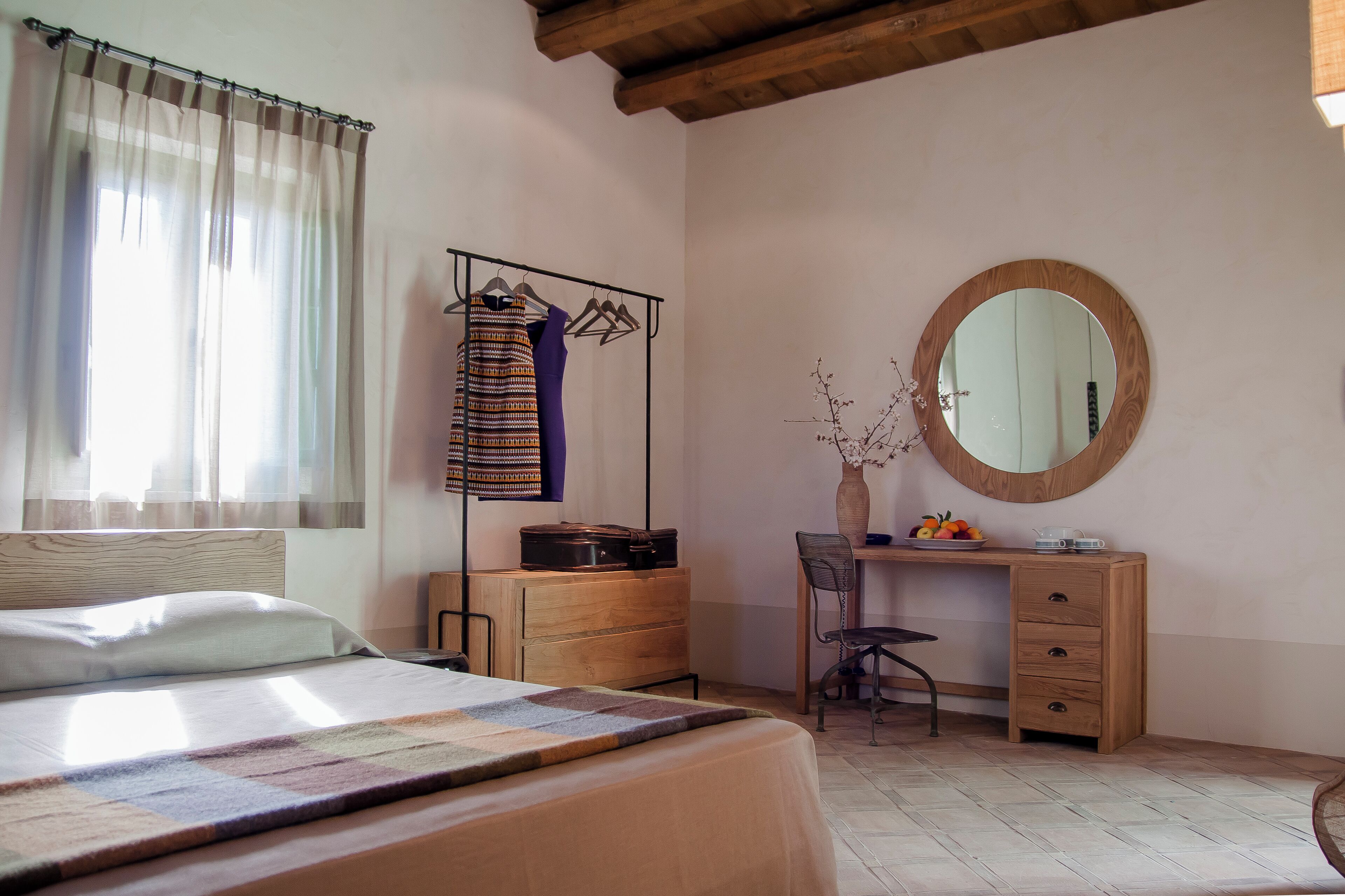 A bedroom with a bed, a wooden dresser, a desk with a chair, a mirror, and a window with curtains.