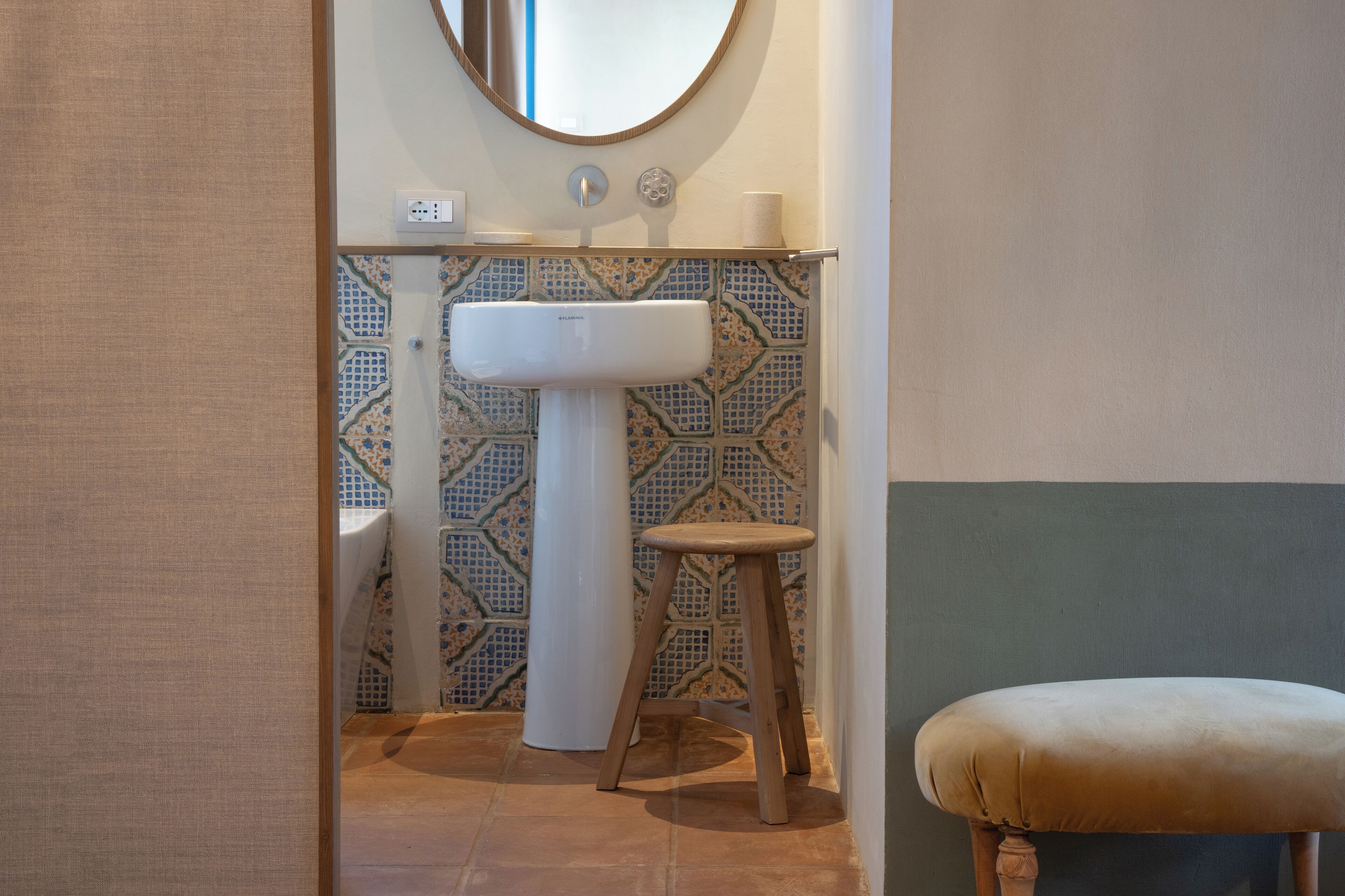 A bathroom with a white pedestal sink, a wooden stool, a round mirror, and patterned tiles.