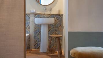 A bathroom with a white pedestal sink, a wooden stool, a round mirror, and patterned tiles.